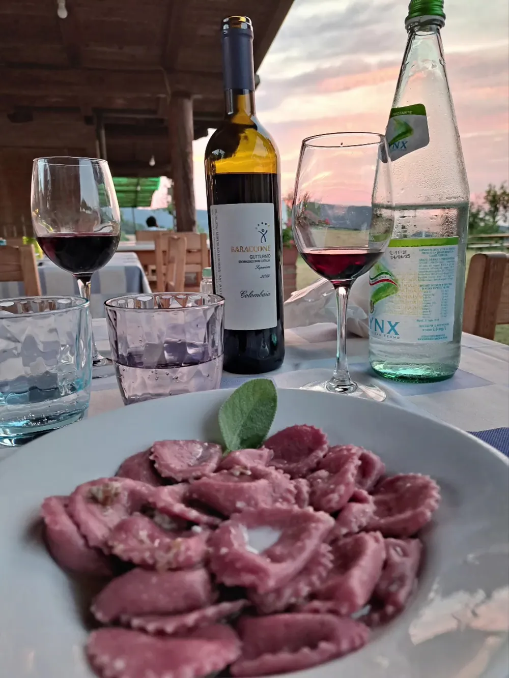 Un piatto di tortelli con la salvia, una bottiglia di Gutturnio e due calici al tramonto.